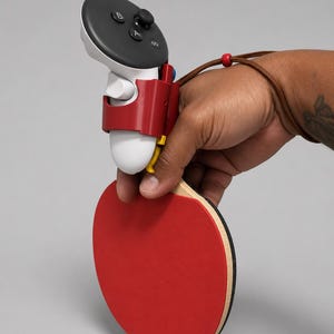 May include: A white and black video game controller attached to a red table tennis paddle. The controller features A, B, and X buttons, a joystick, and a brown leather wrist strap with a red bead. The paddle has a bright red rubber surface.