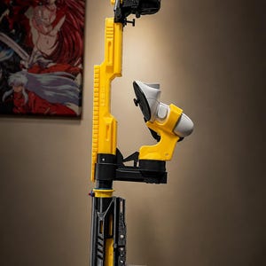 May include: A vertical gaming controller mount featuring a bright yellow rifle-style frame with a black base and stock. The structure holds a white gaming controller in a central cradle and includes a top-mounted attachment for additional hardware.