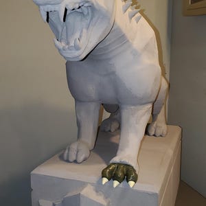 May include: A gray, stylized gargoyle statue with an open mouth, sharp teeth, and red eyes. The statue has pointed ears and a spiky ridge along its back. It stands on a gray, rectangular base, with green and white claws.