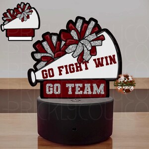 Op de afbeelding: Een witte en rode cheerleader-megafoon met zilveren glitterpompons. De megafoon heeft de tekst "Go Fight Win" en "Go Team" erop. De megafoon staat op een zwarte basis met een schakelaar.