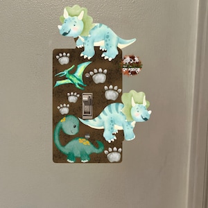 May include: A decorative light switch cover with a brown background and dinosaur illustrations. The design includes a green and blue triceratops, a green dinosaur, and a green pterodactyl. Paw prints are also included in the design.