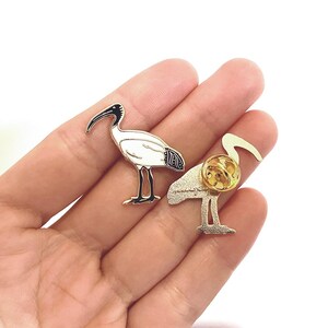 May include: White and black enamel pin of a white ibis bird with a gold finish. The pin is shown from the front and back.