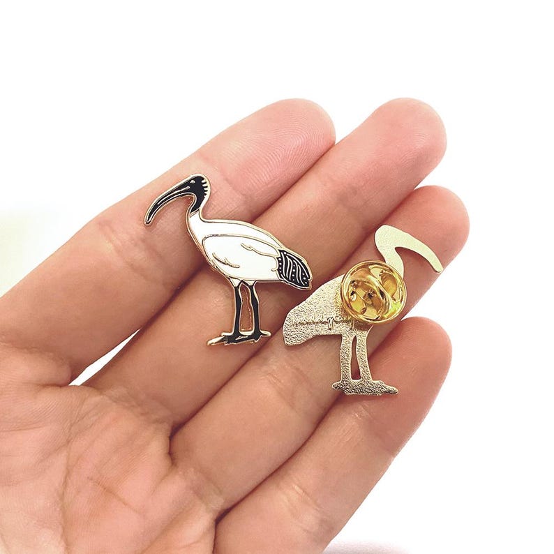 May include: White and black enamel pin of a white ibis bird with a gold finish. The pin is shown from the front and back.