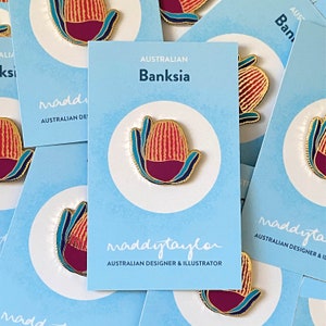 May include: A gold-coloured enamel pin featuring a stylised red and yellow Banksia flower with blue and green leaves. The pin is on a blue background with the text "Australian Banksia" and "maddytaylar Australian Designer & Illustrator".
