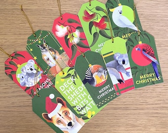 May include: A collection of Christmas gift tags with Australian animal illustrations. The tags feature a kangaroo, koala, cockatoo, and other animals, with red and green backgrounds. Each tag has a gold string for hanging and the words "Merry Christmas" or "Seasons Greetings".