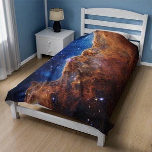 Può includere: Un letto singolo con un copripiumino raffigurante una nebulosa spaziale. Il copripiumino mostra sfumature di blu, arancione e marrone, con numerose stelle. Il telaio del letto è bianco e la stanza ha pareti blu e pavimento in legno.