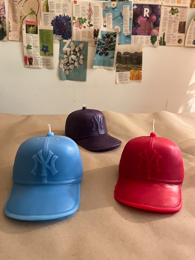 May include: Three baseball cap shaped candles in blue, red, and purple. Each candle has the New York Yankees logo on the front.