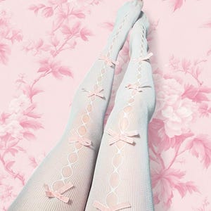 Puede incluir: Medias blancas estampadas con lazos rosas. Las medias presentan un diseño de rombos con pequeños lazos rosas adornando las piernas. El fondo es un estampado floral rosa.