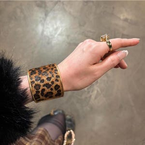 May include: A gold ring with a large, clear gemstone and a wide gold band. A wide cuff bracelet with a leopard print design and gold trim. The bracelet is worn on the wrist.