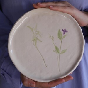 Könnte beinhalten: Ein weißer Keramikteller mit einem gepressten Blumendesign. Der Teller zeigt zwei grüne Pflanzen mit violetten Blüten.