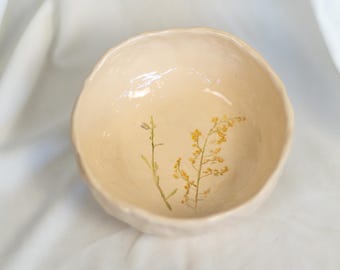 May include: A small, cream-colored ceramic bowl with a textured exterior. The bowl has two pressed dried flowers inside, one with yellow petals and one with white petals.