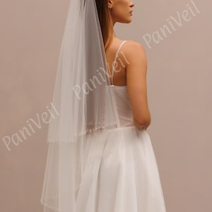 May include: A white bridal veil with a single tier and a beaded edge. The veil is worn by a person with long brown hair.