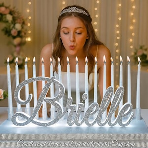 May include: A silver glitter "Brielle" name sign with a white base, surrounded by lit candles. The sign is on a table, with a woman blowing out the candles. The text reads "Font type: Great Vibes on White Base with Silver Theme."