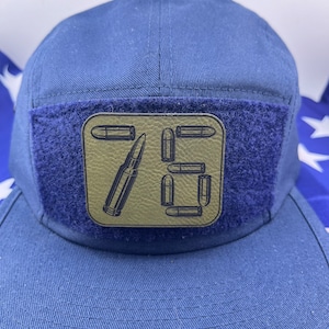 May include: A blue 5-panel hat with a velcro patch featuring a black silhouette of a bullet and the number 75. The patch is on a green background.
