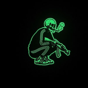 May include: A neon green graphic on a black background depicts a squatting skeleton smoking a cigarette and holding an AK-47. The skeleton wears a tracksuit with three stripes and Adidas sneakers.