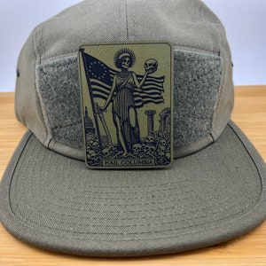 Peut inclure: Une casquette de style militaire verte avec un patch velcro repr&eacute;sentant une illustration en noir et blanc d'une figure squelettique tenant un cr&acirc;ne et un drapeau. Le texte "HAIL COLUMBIA" est imprim&eacute; sous l'image.