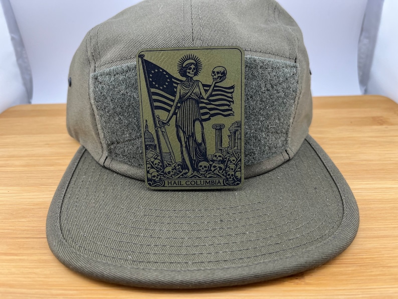 Peut inclure: Une casquette de style militaire verte avec un patch velcro repr&eacute;sentant une illustration en noir et blanc d'une figure squelettique tenant un cr&acirc;ne et un drapeau. Le texte "HAIL COLUMBIA" est imprim&eacute; sous l'image.