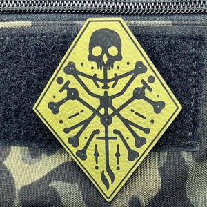 May include: A yellow diamond-shaped patch with a black skull and crossbones design. The skull has a pointed top and a wide, open mouth. The crossbones are made up of four long bones that extend from the skull.
