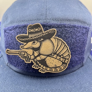 May include: A navy blue baseball cap with a dark blue patch featuring a cartoon armadillo wearing a sombrero and holding a gun. The armadillo is tan with black outlines.