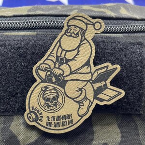 May include: A brown leather patch with a cartoon image of Santa Claus riding a bomb with the text "EGGNOG TO: THE ANTI-HUGGERS FROM: SANTA WITH LOVE".