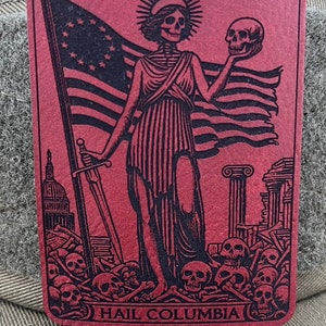 Peut inclure: Un patch rouge avec une image en noir et blanc d'une figure squelettique tenant un cr&acirc;ne et une &eacute;p&eacute;e. La figure se tient devant un drapeau avec des &eacute;toiles et des rayures. La figure se tient sur un tas de cr&acirc;nes et d'os. Le texte "HAIL COLUMBIA" est en bas du patch.