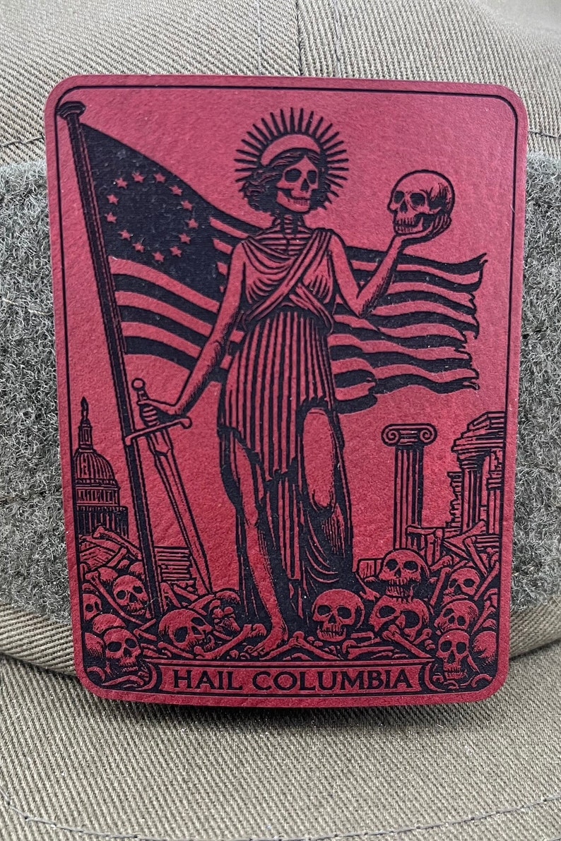 Peut inclure: Un patch rouge avec une image en noir et blanc d'une figure squelettique tenant un cr&acirc;ne et une &eacute;p&eacute;e. La figure se tient devant un drapeau avec des &eacute;toiles et des rayures. La figure se tient sur un tas de cr&acirc;nes et d'os. Le texte "HAIL COLUMBIA" est en bas du patch.