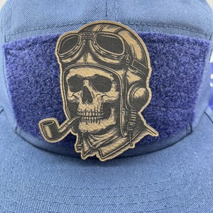 May include: A blue baseball cap with a patch featuring a skull wearing a pilot's helmet and goggles, smoking a pipe. The patch is tan with black details. The cap has a blue velcro patch.