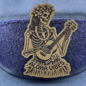 May include: A black and white illustration of a skeleton playing a guitar with a lei of flowers around its neck. The skeleton is wearing a skirt with the text "Aloha Vibes" on it.