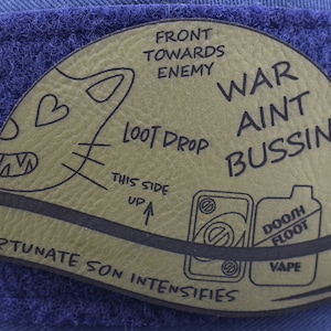 Peut inclure: Un patch en cuir marron avec un visage de chat de dessin animé, le texte "Front towards enemy", "Loot drop", "This side up", "War ain't bussin", et des images d'une bouteille de vape et d'une canette. Le texte "Fortunate son intensifies" est écrit autour du bas du patch.