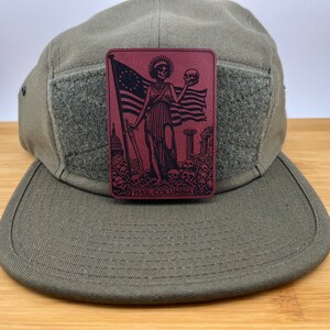 Peut inclure: Un chapeau en toile vert &agrave; 5 panneaux avec un patch velcro sur le devant. Le patch est de couleur bordeaux avec une illustration en noir et blanc d'une figure squelettique tenant un cr&acirc;ne et un drapeau am&eacute;ricain. Le texte "HAIL COLUMBIA" est imprim&eacute; en bas du patch.