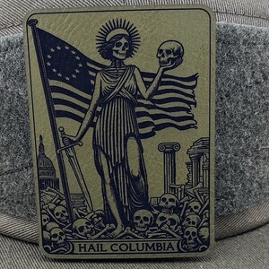 Peut inclure: Une illustration en noir et blanc d'une figure squelettique tenant un cr&acirc;ne et une &eacute;p&eacute;e, debout devant un drapeau am&eacute;ricain en lambeaux. La figure est entour&eacute;e de cr&acirc;nes et d'os. Le texte "HAIL COLUMBIA" est imprim&eacute; en bas de l'image.