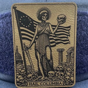 Peut inclure: Une carte dor&eacute;e avec une illustration en noir et blanc d'une figure squelettique tenant un cr&acirc;ne et une &eacute;p&eacute;e. La figure se tient devant un drapeau am&eacute;ricain en lambeaux et un tas d'os. Le texte "HAIL COLUMBIA" est imprim&eacute; en bas de la carte.