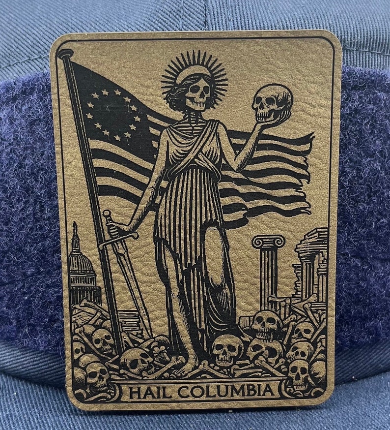 Peut inclure: Une carte dor&eacute;e avec une illustration en noir et blanc d'une figure squelettique tenant un cr&acirc;ne et une &eacute;p&eacute;e. La figure se tient devant un drapeau am&eacute;ricain en lambeaux et un tas d'os. Le texte "HAIL COLUMBIA" est imprim&eacute; en bas de la carte.
