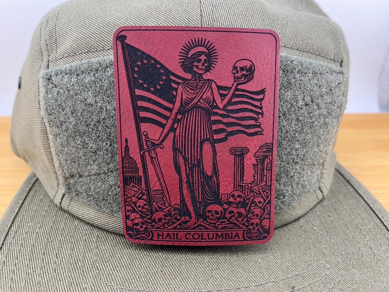 Peut inclure: Un patch rouge avec une image en noir et blanc d'une figure squelettique tenant un cr&acirc;ne. La figure se tient devant un drapeau am&eacute;ricain en lambeaux et une &eacute;p&eacute;e. Le texte "HAIL COLUMBIA" est imprim&eacute; en bas du patch.