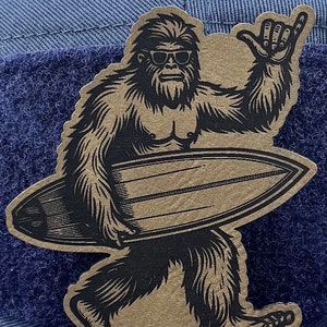 May include: A brown and black illustrated patch of a Bigfoot character wearing sunglasses and holding a surfboard with one hand raised in a 'hang loose' gesture.
