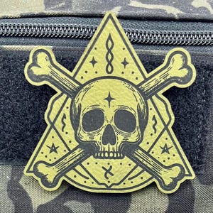 May include: A yellow triangle patch with a black skull and crossbones design. The skull has a five-pointed star on its forehead. The patch has a geometric pattern with stars and a symbol that looks like an infinity sign.