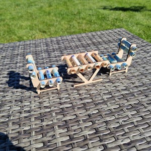 May include: A miniature furniture set consisting of a table and two chairs constructed from light-colored wood. The seats and tabletop are formed by multiple rolled-up paper currency bills in shades of blue, green, and white, secured within the frames.