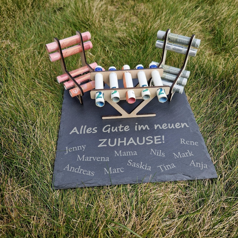 May include: A dark gray slate plaque engraved with the text Alles Gute im neuen ZUHAUSE and various names, featuring a wooden structure holding rolled paper currency. The display includes two chair-like wooden frames and a central rack for bills.