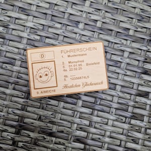 Könnte beinhalten: Holzplakette mit deutschem Text, darunter "FÜHRERSCHEIN", ein Name und ein Geburtsdatum. Die Plakette zeigt ein Cartoon-Gesicht und ist rechteckig mit abgerundeten Ecken.