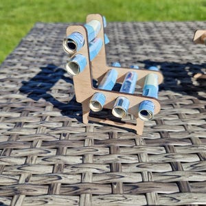 May include: A small, light-colored wooden chair structure featuring several blue paper rolls inserted into circular cutouts along the backrest and seat. The frame is constructed from thin, laser-cut wood panels, creating a miniature furniture piece.