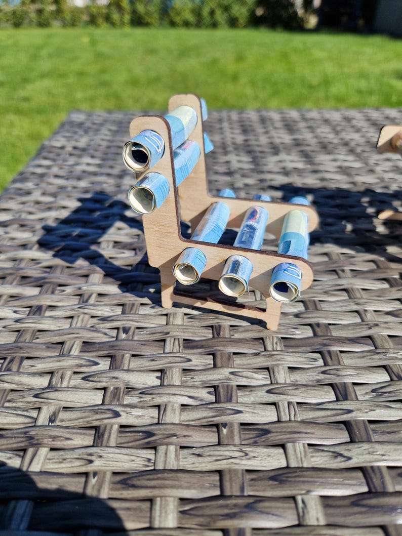 May include: A small, light-colored wooden chair structure featuring several blue paper rolls inserted into circular cutouts along the backrest and seat. The frame is constructed from thin, laser-cut wood panels, creating a miniature furniture piece.