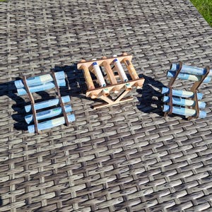 May include: Three wooden racks designed to hold rolled paper currency. Each rack features a light brown finish and a structural frame with multiple horizontal slots. The racks are filled with several light blue rolled paper bills.