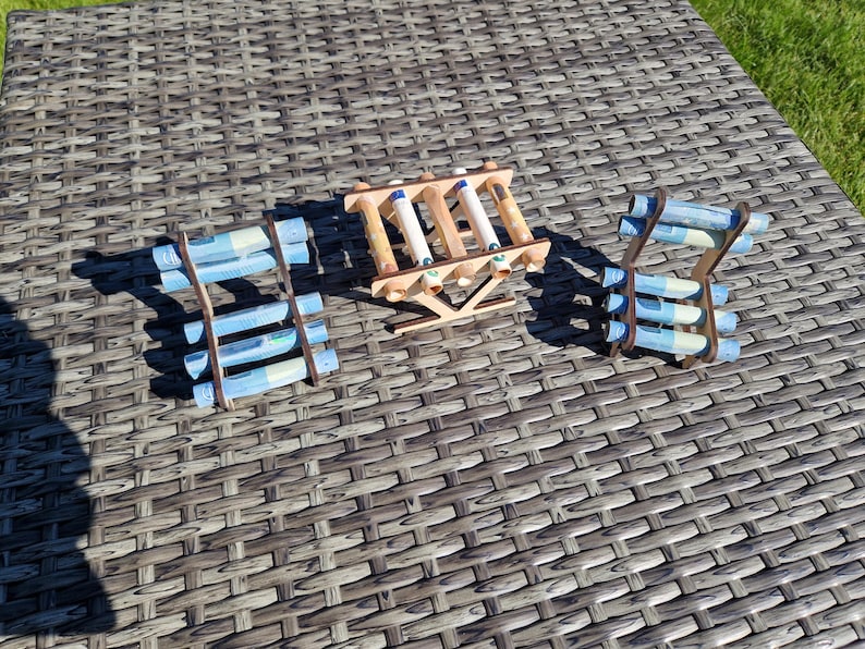 May include: Three wooden racks designed to hold rolled paper currency. Each rack features a light brown finish and a structural frame with multiple horizontal slots. The racks are filled with several light blue rolled paper bills.