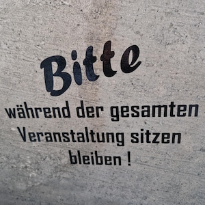 Könnte beinhalten: Ein Schild an einer Betonwand mit schwarzem Text, der "Bitte während der gesamten Veranstaltung sitzen bleiben!" lautet, was übersetzt "Bitte bleiben Sie während der gesamten Veranstaltung sitzen!" bedeutet.
