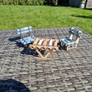 May include: A miniature set consisting of a small table and two chairs constructed from light-colored wood. The table and chairs feature decorative accents made from rolled-up light blue and white paper currency notes tucked into the wooden frame.