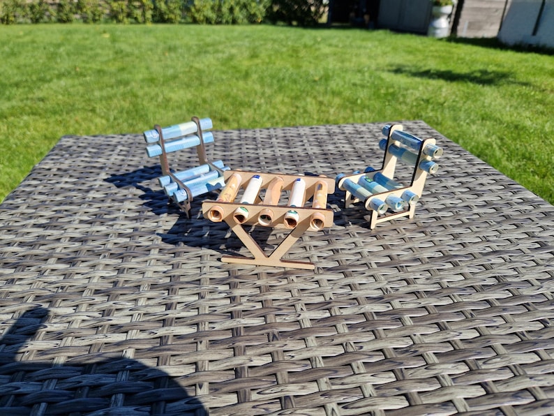 May include: A miniature set consisting of a small table and two chairs constructed from light-colored wood. The table and chairs feature decorative accents made from rolled-up light blue and white paper currency notes tucked into the wooden frame.