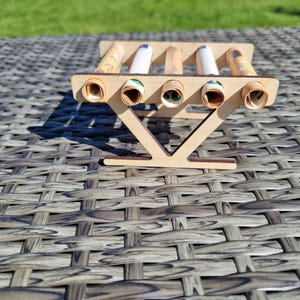 May include: A light-colored wooden stand featuring five horizontal circular cutouts, each holding a tightly rolled paper banknote. The structure is supported by a V-shaped base, providing a stable platform for the display of the rolled currency items.