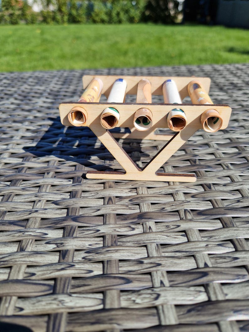 May include: A light-colored wooden stand featuring five horizontal circular cutouts, each holding a tightly rolled paper banknote. The structure is supported by a V-shaped base, providing a stable platform for the display of the rolled currency items.