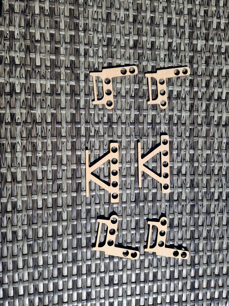 May include: Six light-colored wooden laser-cut components arranged in three pairs. The top and bottom pairs are L-shaped with two circular holes on one arm and three on the other. The middle pair features a triangular brace with five holes.