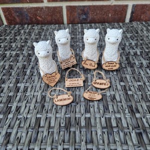 Könnte beinhalten: Eine Ansammlung weißer Alpaka-Figuren mit Holzschildern. Die Schilder tragen verschiedene deutsche Sätze, darunter "Lächle!" und "Für Dich". Die Figuren sind auf einer grauen, gewebten Oberfläche angeordnet.